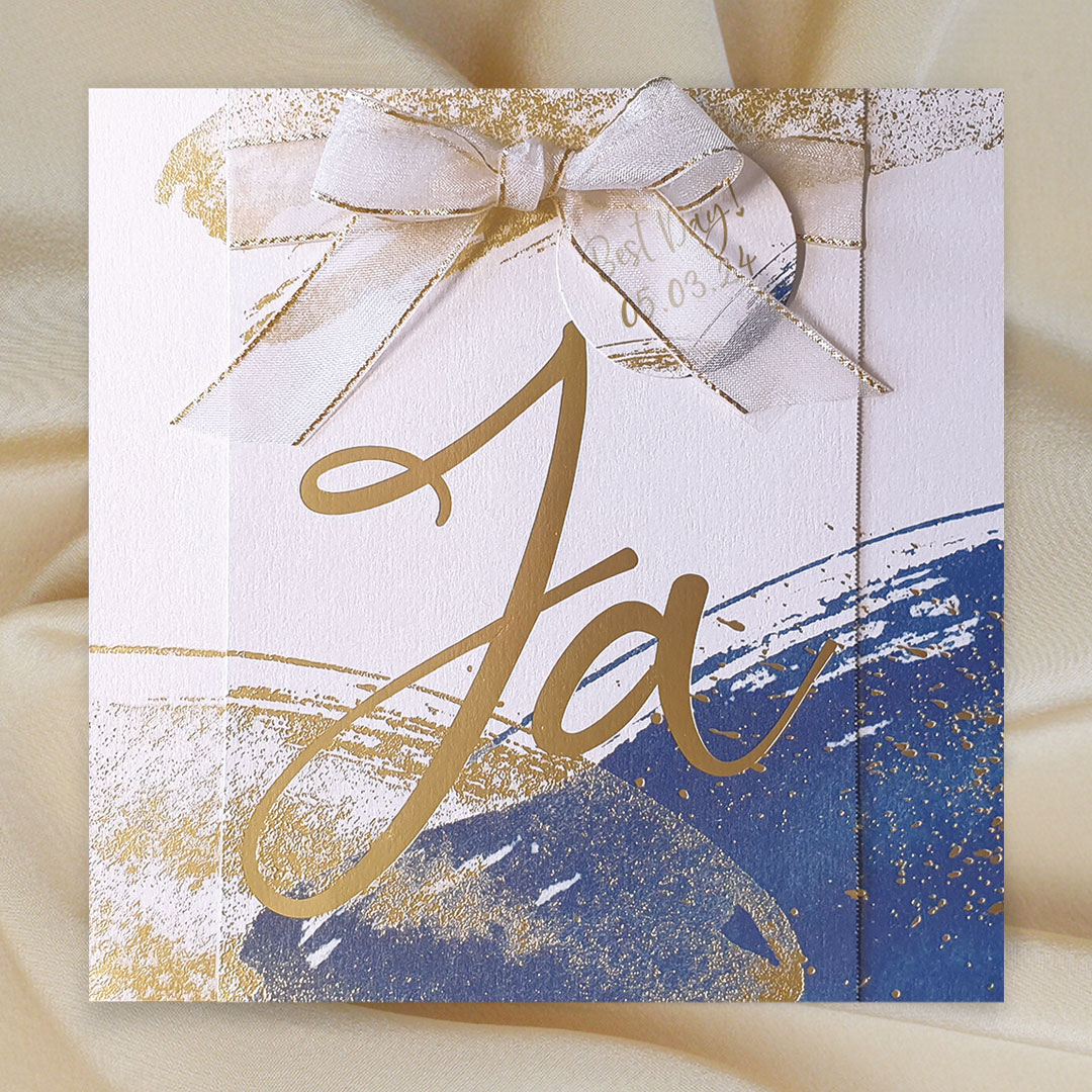 Vergrößerte Abbildung Weiße Hochzeitseinladungen mit blauen und goldenen Akzenten auf goldenem Stoff liegend.